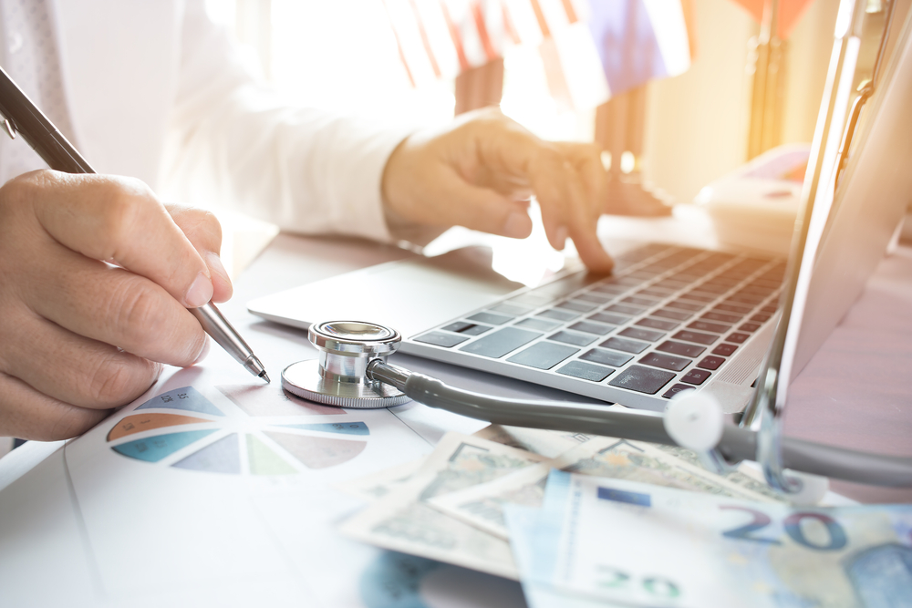 Doctor,Working,On,Laptop,Computer,With,Report,Analysis,And,Money
